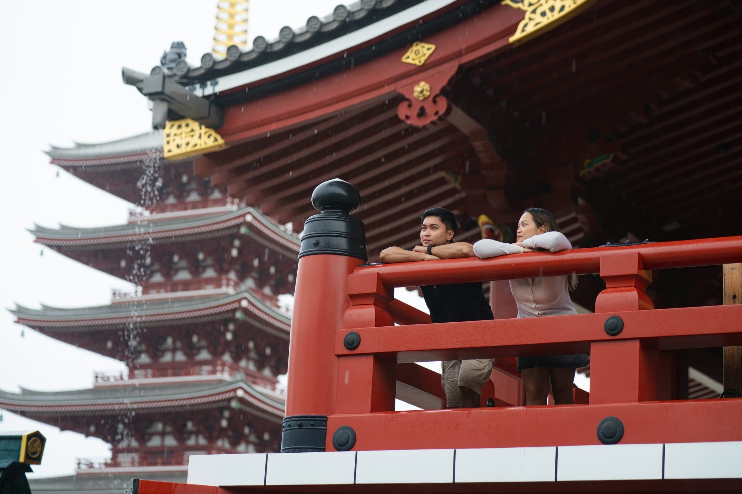 A couple leaning on a red balcony rail with the five-storey pagoda behind