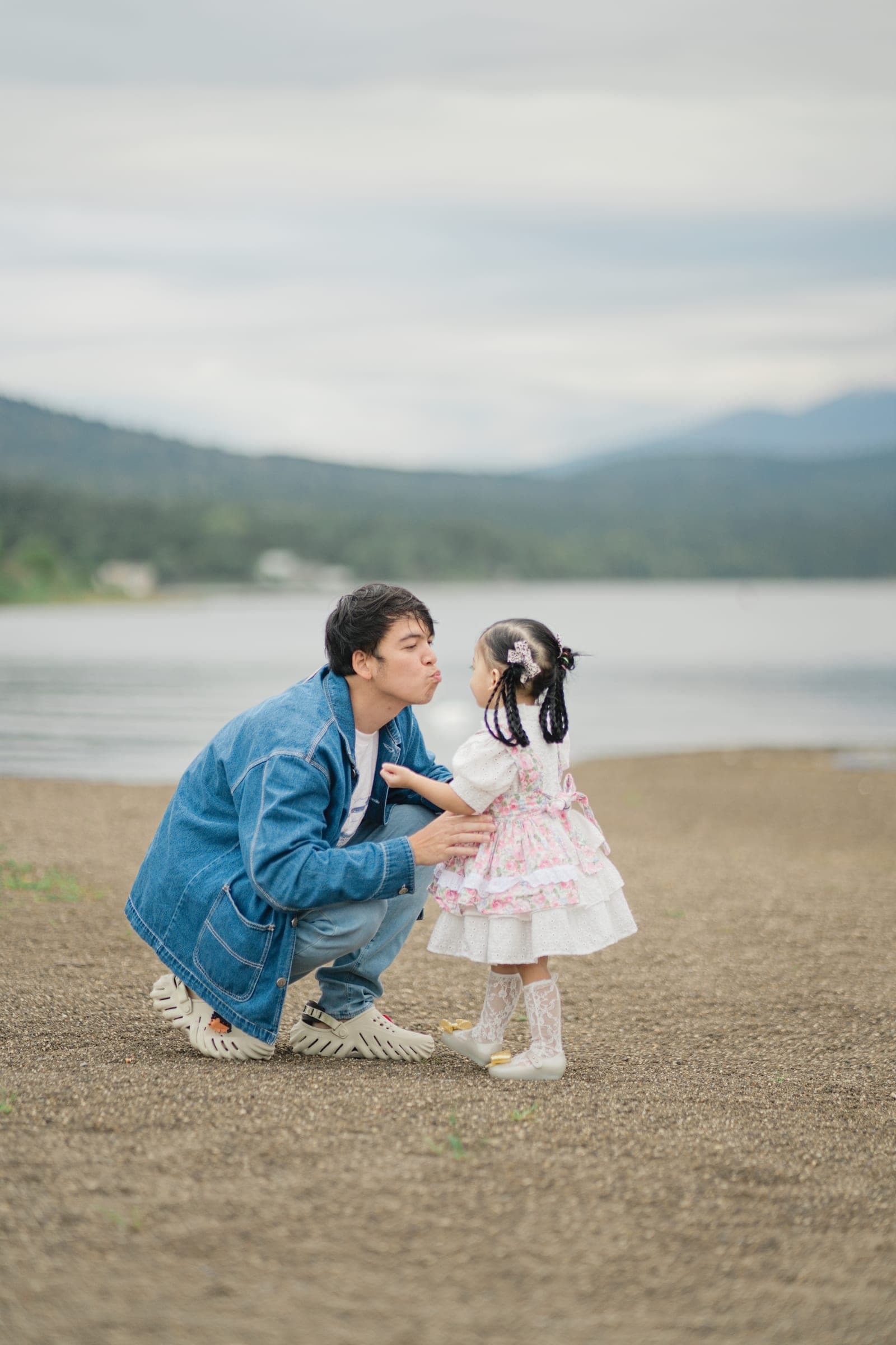 A father crouching at the water's edge, pulling a face at his small daughter